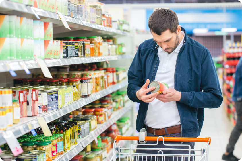 At the Supermarket: Handsome Man Uses Smartphone and Looks at Nutritional Value of the Canned Goods. He's Standing with Shopping Cart in Canned Goods Section.